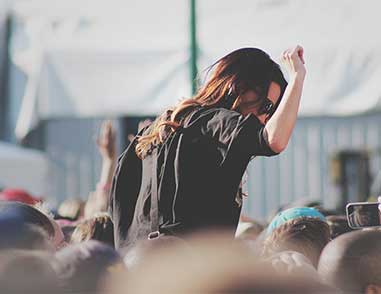 Парень с длинными волосами и в солнзезащитных очках, подняв правую руку наверх и у кого то на плечах среди толпы фанатов