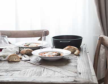 На кухонном столе, которая стоит возле окна сковорода, тарелки с едой, бокалы с напитком, ложка и куски хлеба