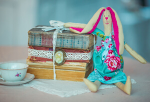 На столе самодельная тряпочная кукла, стопка книг и чашечка с блюдцем